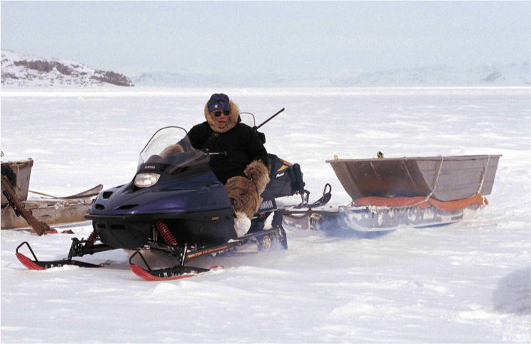 A man sits on a snowmobile with a sled hitched behind it, the flat snow stretching out behind him into the distance